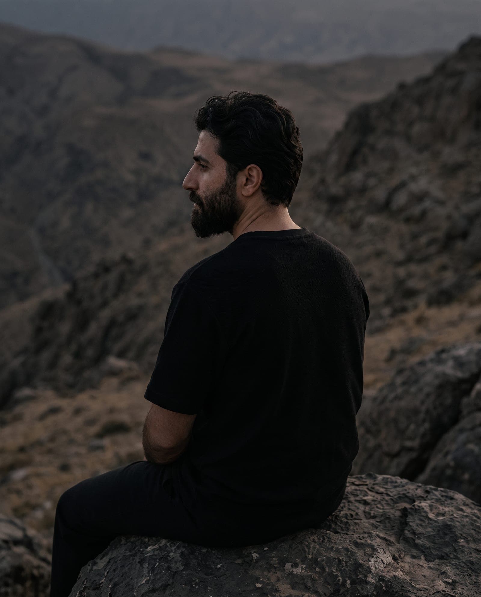 A man in a plain black heavyweight t-shirt sitting on a boulder above a mountain valley at dusk